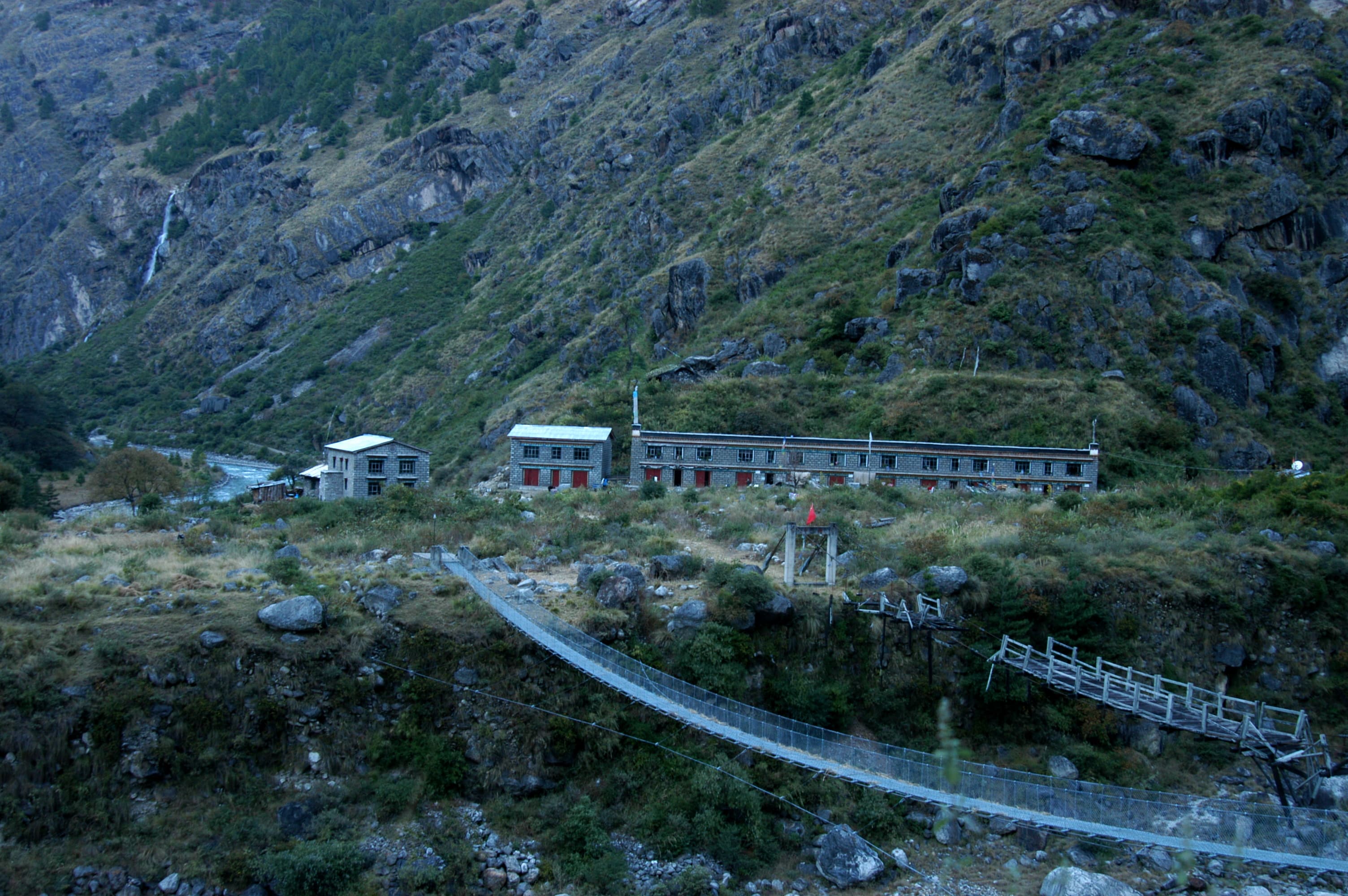 A suspension bridge and stone buildings along a rugged mountain slope in the Bhote Kosi river basin.