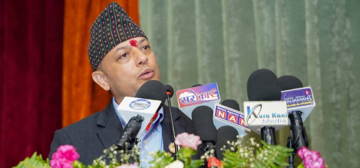 Dol Prasad Aryal delivering an address into several microphones behind floral arrangements.