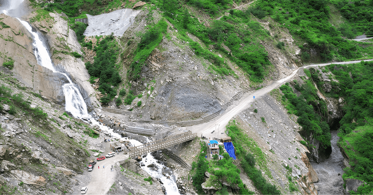A narrow mountain road crosses a stream beside a steep waterfall and rocky cliffs.