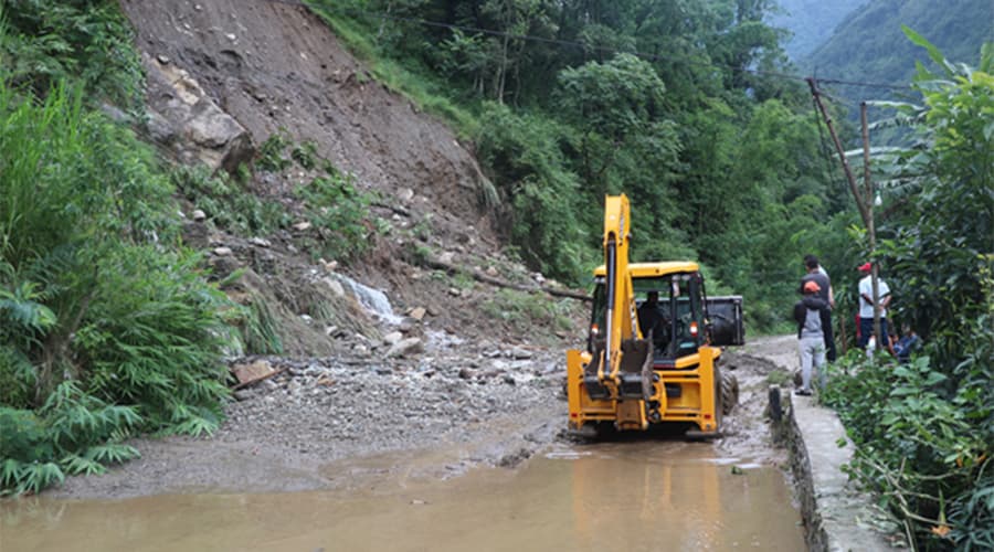 A yellow backhoe loader clears mud and landslide debris from a waterlogged mountain road.