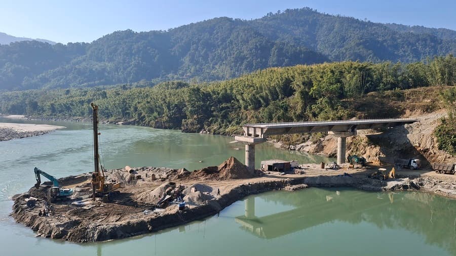 Construction site of an incomplete motorable bridge across the turquoise waters of the Sunkoshi River, with exposed pillars and machinery.