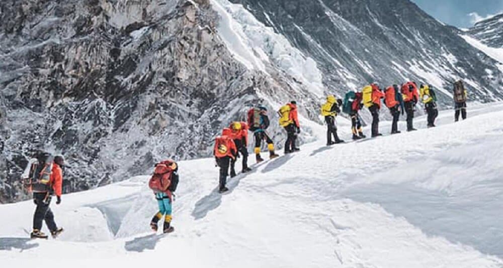 A line of mountaineers wearing climbing gear ascends a snowy mountain slope in the Himalayas.