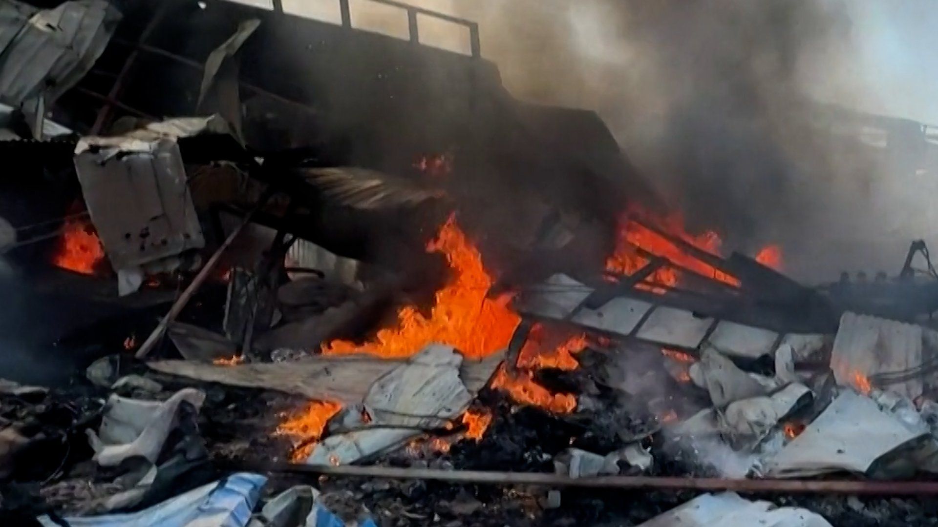 Flames consume destroyed structures and corrugated metal near a bridge following a military airstrike in Myanmar.