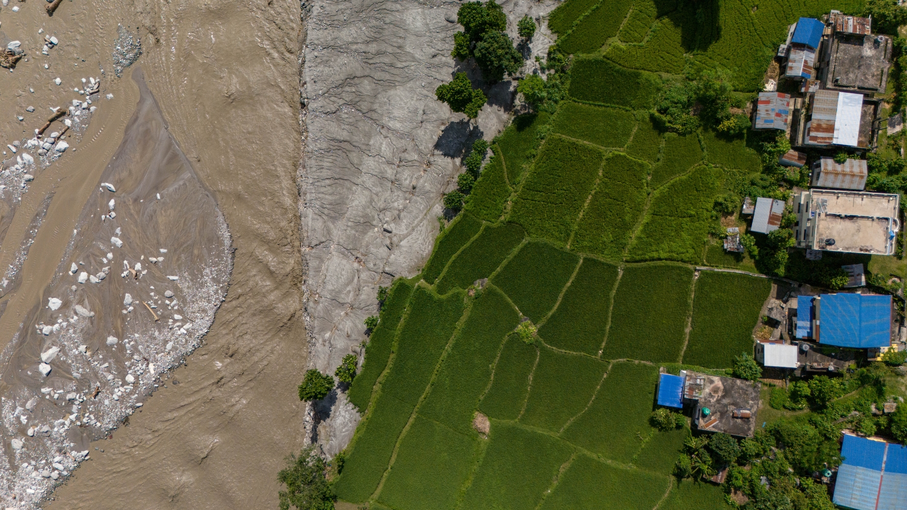 Aerial view of flood sediment and a turbid river encroaching upon green terraced fields and residential homes.